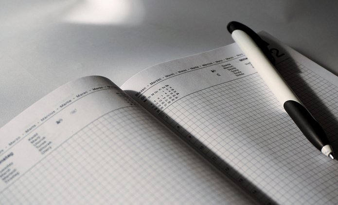 Na zdjęciu widać otwarty notes lub planer leżący na jasnej powierzchni. Obie strony mają delikatną kratkę do pisania. U góry strony widoczny jest nagłówek z nazwami miesięcy w różnych językach, m.in. „Březen”, „Marec”, „März”, „Marzo”, co wskazuje na marzec. Na prawej stronie notesu, ukośnie, leży biało-czarny długopis. Światło pada z lewej strony, tworząc wyraźne cienie długopisu i zagiętej kartki. Całość ma spokojny, minimalistyczny charakter i sugeruje planowanie lub notowanie.