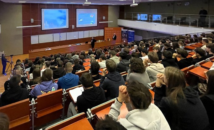 Duża sala wykładowa wypełniona uczniami i uczestnikami wydarzenia. Wszyscy siedzą w rzędach skierowanych w stronę sceny, gdzie prowadzący prezentuje materiał wyświetlany na dwóch dużych ekranach. Widać jasne oświetlenie, drewniane ławki i szeroką przestrzeń audytorium. Uczestnicy uważnie śledzą przebieg wykładu.