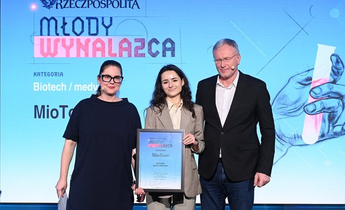 Na zdjęciu widać trzy osoby stojące na scenie podczas uroczystej gali. W centrum znajduje się młoda kobieta ubrana w jasny garnitur, trzymająca w dłoniach oprawiony dyplom. Towarzyszą jej dwie osoby dorosłe — kobieta po lewej stronie i mężczyzna po prawej — ubrane w ciemne, eleganckie stroje. Wszyscy stoją przodem do obiektywu i pozują do zdjęcia. W tle widoczny jest duży ekran z napisem „Rzeczpospolita – Młody Wynalazca” oraz informacją o kategorii „Biotech / medycyna”. Scena jest profesjonalnie oświetlona, a całość ma oficjalny, galowy charakter.