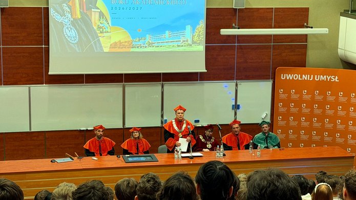 Widok auli podczas uroczystej inauguracji roku akademickiego 2026/2027 na Wydziale Fizyki i Informatyki Stosowanej UŁ. Przy stole prezydialnym siedzą przedstawiciele władz uczelni w togach i biretach, a na środku przemawia Dziekan Wydziału. Na ekranie wyświetla się grafika inauguracji, a na pierwszym planie widać zgromadzonych studentów i gości.