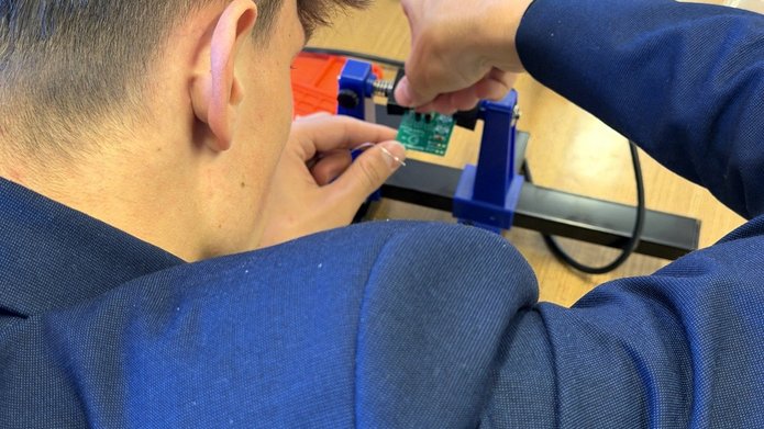 Uczeń podczas warsztatów z elektroniki przytrzymuje płytkę drukowaną z układem elektronicznym, pracując pod kierunkiem prowadzącego. Zajęcia odbywają się przy stanowisku do lutowania.